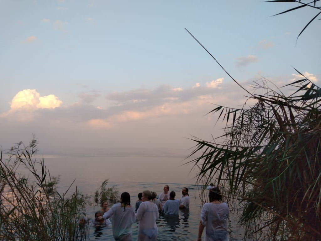 Группа людей в белых рубаках идет по пояс в озере, окруженном камышами